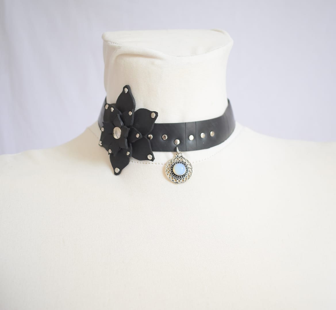 Recyled Jewellery: A white mannequin displays a Flower XL Choker Moon Stone adorned with silver studs. The choker features a black leather flower with a silver center on one side and a small, round pendant with a light blue moon stone in an ornate silver setting at the front. @ Reblack Shop