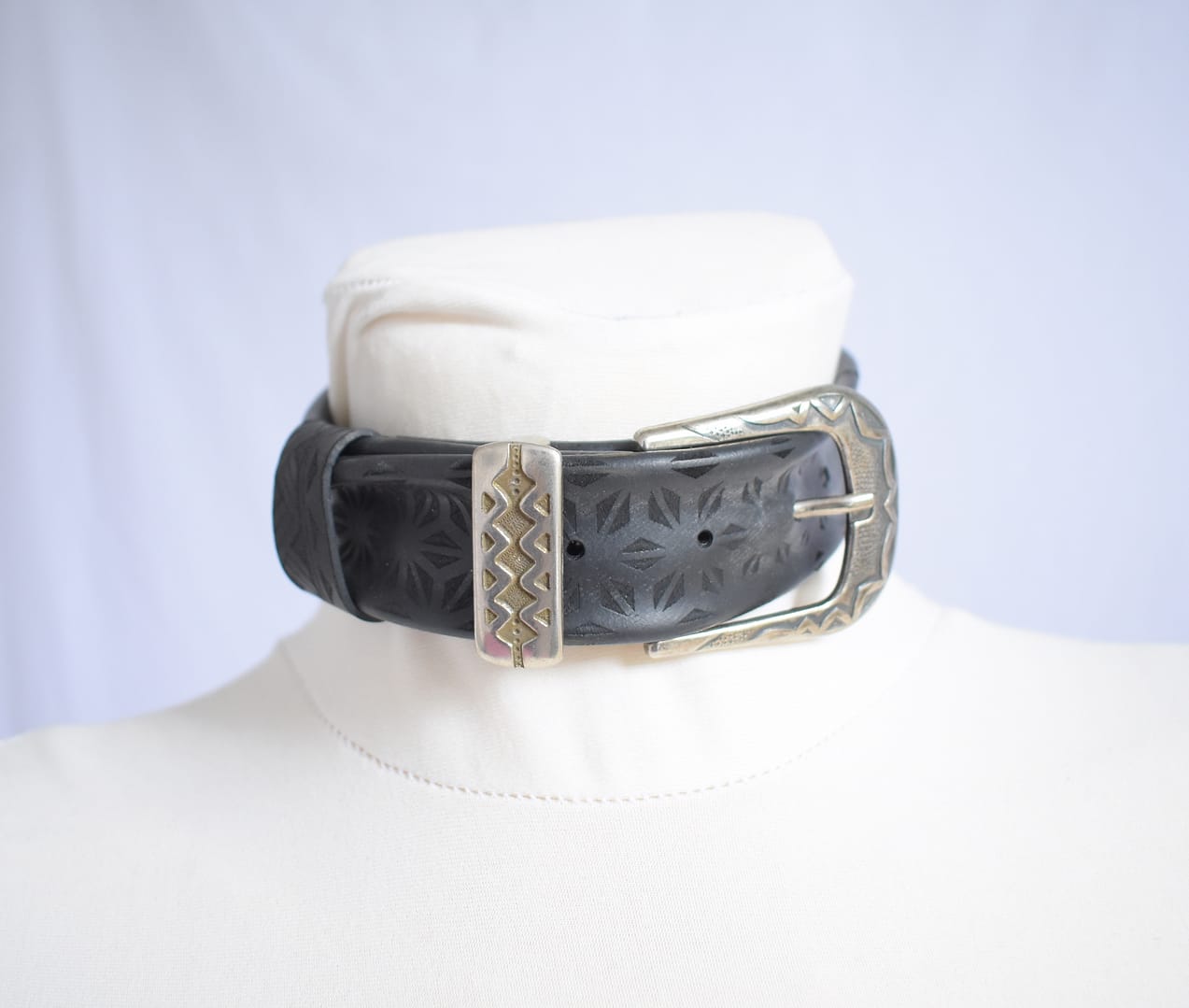 Recyled Jewellery: A black leather Belt Choker with a large, ornate silver buckle rests around the neck of a white mannequin. The Belt Choker features embossed geometric patterns, adding texture to its design, resembling the intricate details of a finely crafted belt. The background is plain and light-colored. @ Reblack Shop