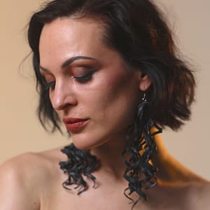 Recyled Jewellery: A woman with short, wavy black hair and dramatic makeup gazes downward. She wears Curly Earrings XL that curl elegantly, enhancing her contemplative expression. The background is a soft, warm gradient. @ Reblack Shop