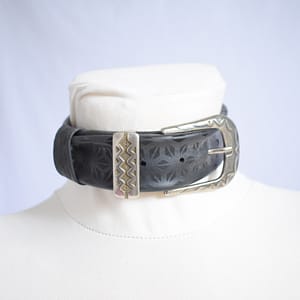 Recyled Jewellery: A black leather Belt Choker with a large, ornate silver buckle rests around the neck of a white mannequin. The Belt Choker features embossed geometric patterns, adding texture to its design, resembling the intricate details of a finely crafted belt. The background is plain and light-colored. @ Reblack Shop