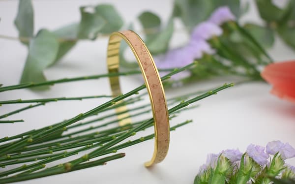 Recyled Jewellery: A golden bracelet with etchings, crafted from recycled jewellery, resting on a sprig of pine needles, surrounded by colorful flowers and green leaves on a white background. @ Reblack Shop
