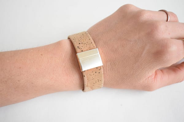 Recyled Jewellery: A close-up of a person's wrist wearing a simple beige wristband made from recycled jewellery with a rectangular silver buckle against a white background. @ Reblack Shop