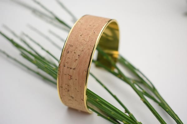 Recyled Jewellery: A golden bracelet with a cork texture, crafted from recycled jewellery, displayed on a white surface, partially laying on green, needle-like leaves. @ Reblack Shop