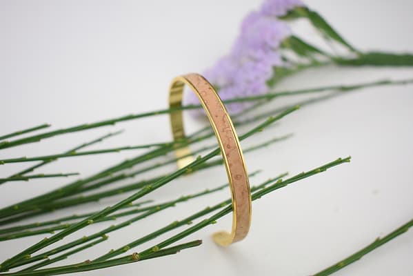 Recyled Jewellery: A golden bracelet crafted from recycled jewellery, with engraved symbols, foregrounded against a backdrop of slender green stems and purple flowers. @ Reblack Shop