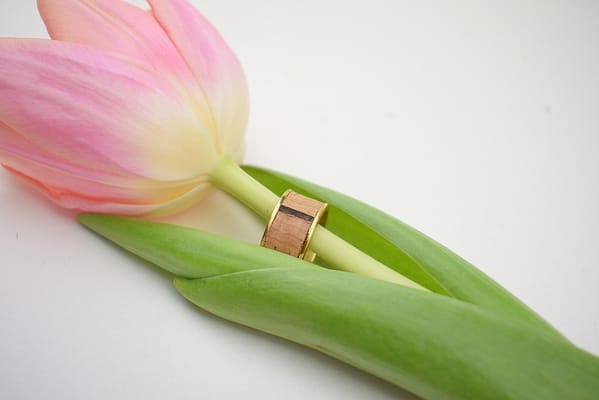 Recyled Jewellery: A close-up image of a soft pink tulip with a recycled gold wedding band placed around one of its green stems on a white background. @ Reblack Shop