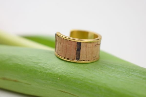 Recyled Jewellery: A close-up photo of a wooden ring with a natural, grainy texture and a golden inner band, crafted from recycled jewellery, resting on a green leaf against a white background. @ Reblack Shop