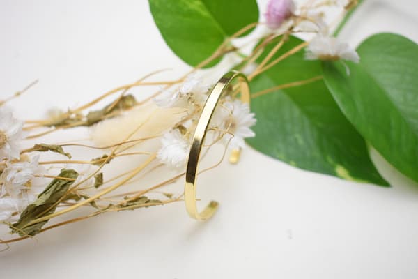 Recyled Jewellery: A golden bracelet crafted from recycled jewellery, surrounded by dried white flowers and fresh green leaves on a white background. @ Reblack Shop