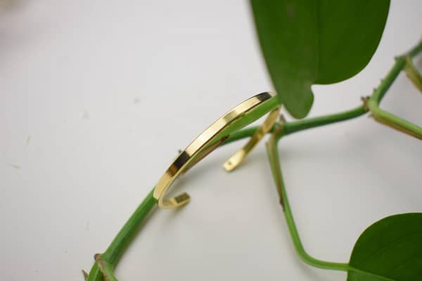 Recyled Jewellery: A golden bracelet made from recycled jewellery resting on a green vine with broad leaves, set against a white background. The focus is on the shiny surface of the bracelet. @ Reblack Shop