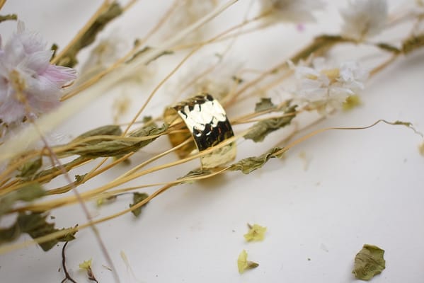 Recyled Jewellery: A golden ring made from recycled jewellery surrounded by delicate dried white flowers and slender stems scattered on a white background. @ Reblack Shop