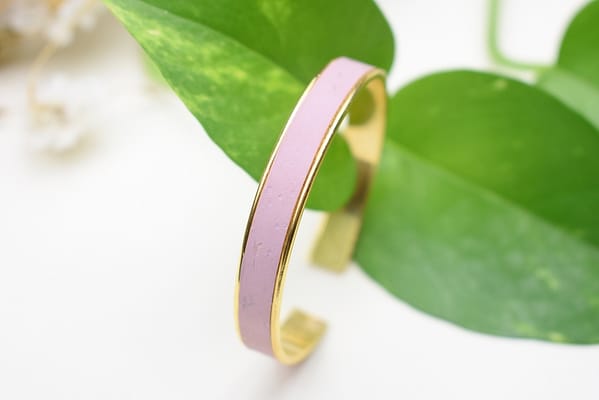 Recyled Jewellery: A close-up of a recycled lavender and gold bangle bracelet resting on a surface, partially surrounded by vibrant green leaves. The background is softly blurred with hints of white. @ Reblack Shop