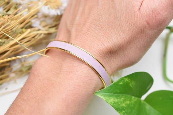 Recyled Jewellery: A close-up image of a person’s wrist wearing a simple recycled jewellery bangle, with a background of dried flowers and green leaves. The bangle is white with a thin golden line. @ Reblack Shop