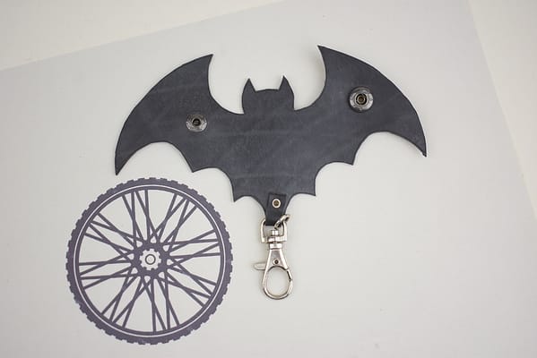 Recyled Jewellery: A bat-shaped keychain in black with clasps and a recycled jewellery-inspired circular patterned segment, lying on a white surface. @ Reblack Shop