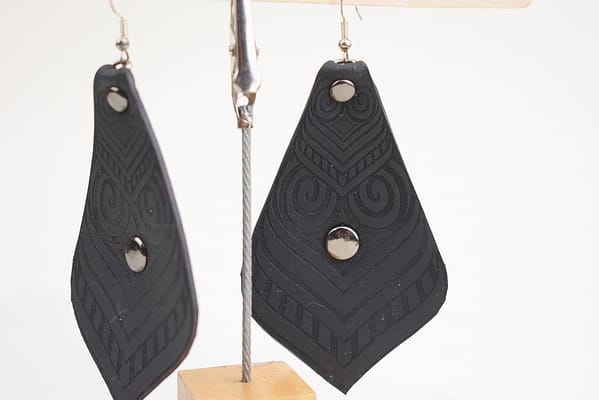 Recyled Jewellery: A pair of black triangular dangling earrings with geometric patterns and a central circular cutout, crafted from upcycled materials, displayed on a wooden stand against a white background. @ Reblack Shop