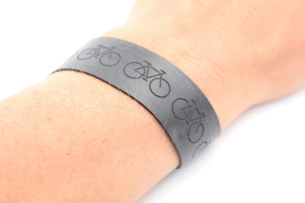 Recyled Jewellery: Close-up of a wrist wearing a black rubber Bicycle Bracelet, eco-friendly jewelry featuring an embossed design of multiple white bicycle symbols. The background is a plain white surface. @ Reblack Shop