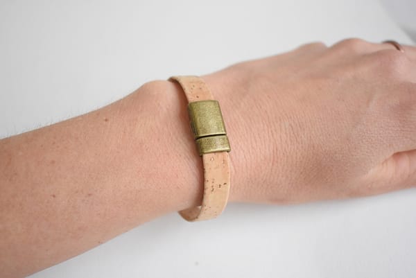 Recyled Jewellery: A close-up image of a person's wrist wearing a simple beige recycled cork bracelet with a metallic gold clasp, set against a plain white background. @ Reblack Shop
