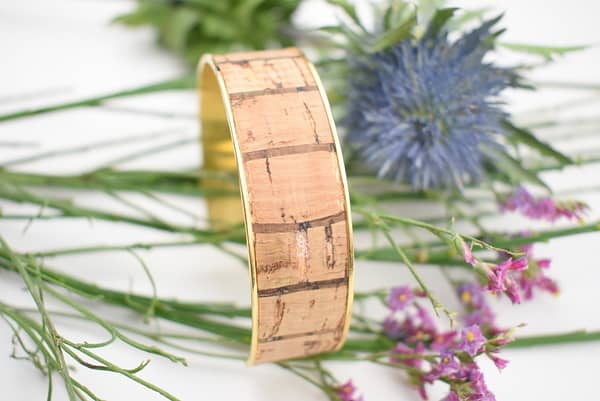 Recyled Jewellery: A recycled cork bracelet on a white background surrounded by green stems and purple and blue flowers, emphasizing eco-friendly fashion. @ Reblack Shop