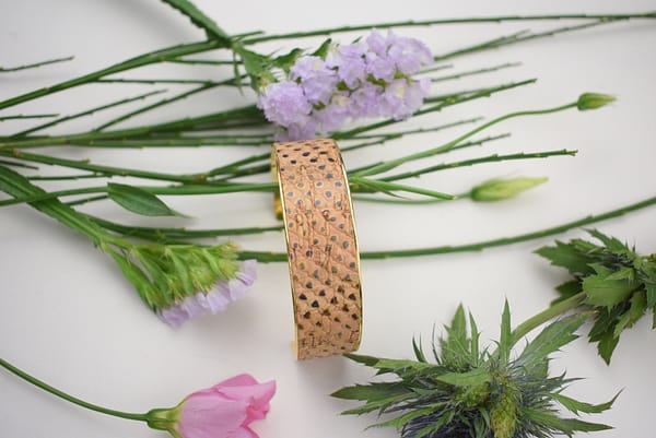 Recyled Jewellery: A recycled brass bracelet with a pattern of small holes lies on a white surface surrounded by green stems and various flowers, including purple and pink blooms. @ Reblack Shop