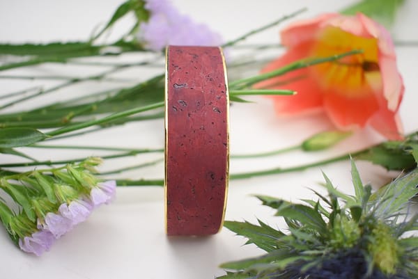 Recyled Jewellery: A close-up of a red bracelet made from recycled jewellery with a textured surface, displayed among a variety of colorful flowers and green leaves on a white background. @ Reblack Shop