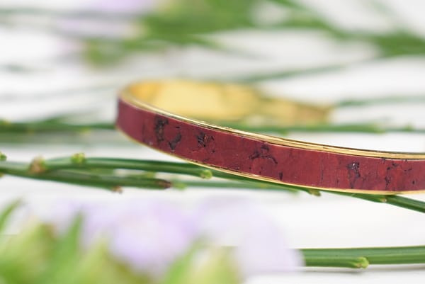 Recyled Jewellery: Close-up of a red recycled bangle with black speckles, surrounded by green plant stems and soft purple flowers in the background. The focus is on the detailed texture of the bangle. @ Reblack Shop