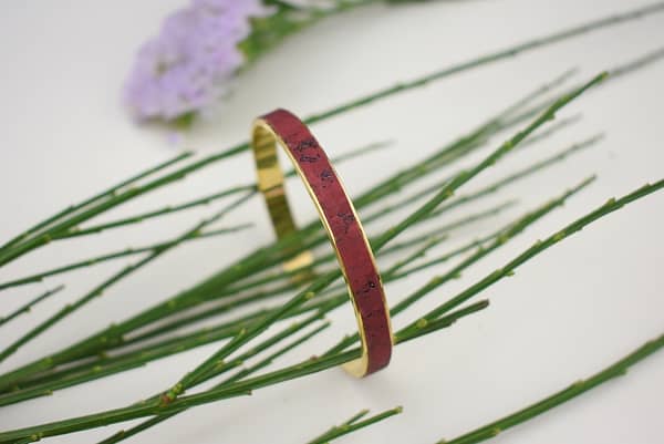 Recyled Jewellery: A recycled copper bracelet with subtle engravings laying on a bed of green, needle-like leaves with a purple flower blurred in the background. @ Reblack Shop