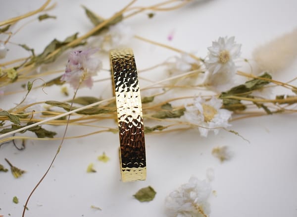Recyled Jewellery: A textured recycled gold bracelet stands upright surrounded by delicate, dried white and pink flowers on a white background. @ Reblack Shop