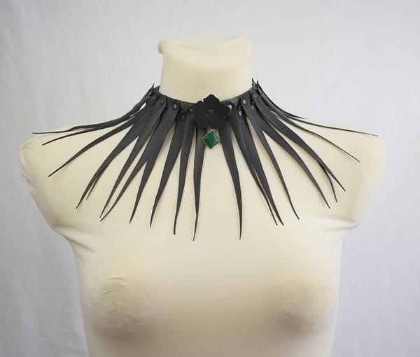 Recyled Jewellery: A black choker necklace with multiple elongated spikes radiating outward is displayed on a white mannequin. Featuring a green gemstone pendant at the center, this AUTO-DRAFT choker stands out beautifully against the plain white background. @ Reblack Shop