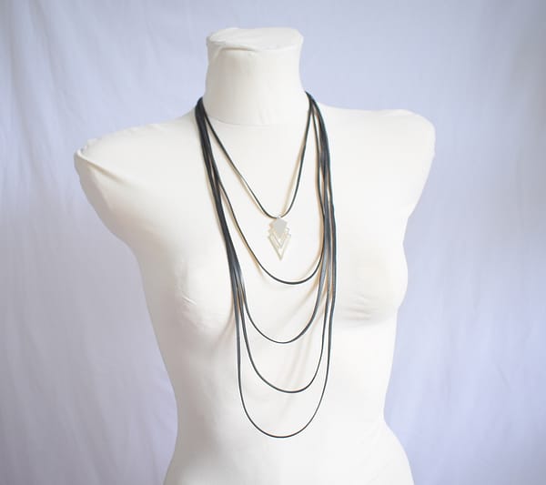 Recyled Jewellery: A white mannequin torso adorned with multiple layered black necklaces of varying lengths gleams charmingly. One striking piece of jewelry, the Step to Step Big Necklace, features a geometric pendant with a triangular design. The background is a plain white fabric, ensuring all focus remains on the elegant showcase. @ Reblack Shop