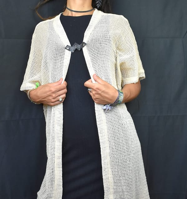 Recyled Jewellery: A woman standing with her hands slightly clutching the edges of her long, knitted white cardigan, over a dark dress adorned with eco-friendly jewelry, against a black background. @ Reblack Shop