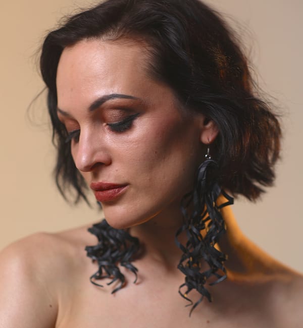 Recyled Jewellery: A woman with short, wavy black hair and dramatic makeup gazes downward. She wears Curly Earrings XL that curl elegantly, enhancing her contemplative expression. The background is a soft, warm gradient. @ Reblack Shop