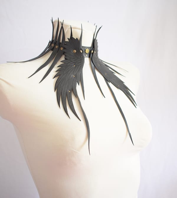 Recyled Jewellery: A white mannequin is adorned with an artistic, black feathered choker necklace from the Dark Angel Jewelry collection. The Dark Angel Choker Brass features long, slender feather-like structures extending outward from a central point, adding a dramatic flair. The background is plain and white, emphasizing the necklace's design. @ Reblack Shop