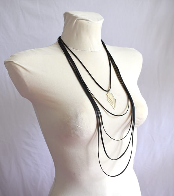 Recyled Jewellery: A white mannequin torso displays a multi-layered, big black necklace with a geometric silver pendant. The Step to Step Big Necklace features several strands of varying lengths, creating an elegant cascade. Set against a plain white background, this piece is perfect for those who love bold statement pieces. @ Reblack Shop