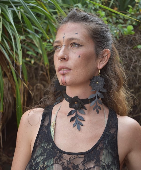 Recyled Jewellery: A woman with star and line face tattoos, wearing a sheer black lace top, large black floral earrings, and a matching choker with leaf accents, stands in front of lush greenery. With her hair pulled back and a nose ring glinting subtly, she exudes an auto-draft allure. @ Reblack Shop