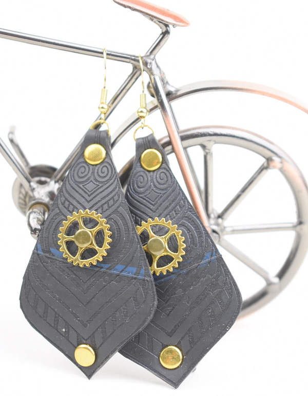Recyled Jewellery: A pair of black earrings made from recycled jewellery, with intricate patterns and small golden gears, displayed against a blurred background featuring a miniature bicycle model. @ Reblack Shop
