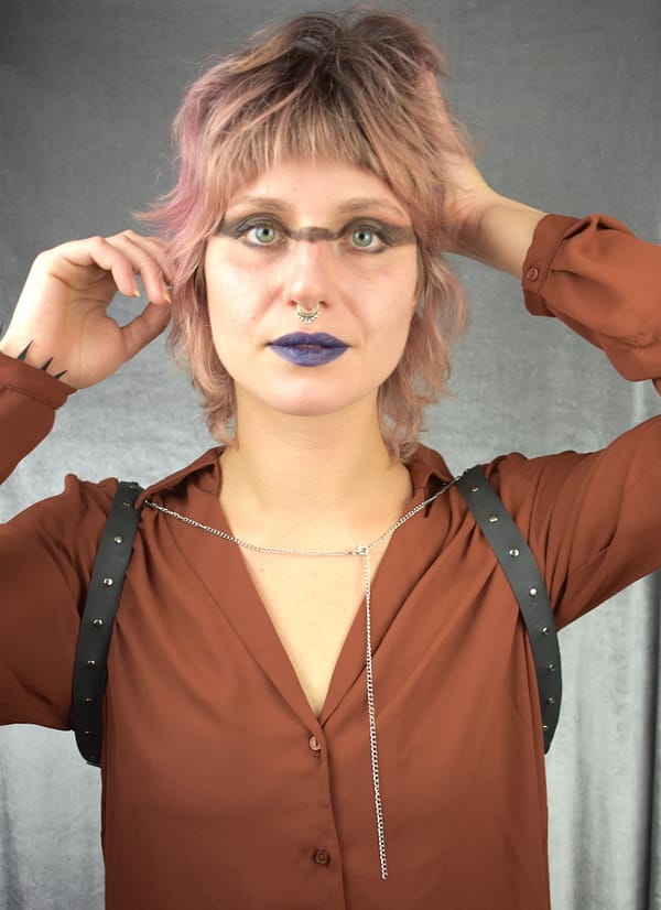 Recyled Jewellery: A woman with pink hair poses with her hands on her head, wearing a brown blouse, the Celina Harness, and blue lipstick. She has an eco-friendly jewelry septum nose ring and looks directly at @ Reblack Shop