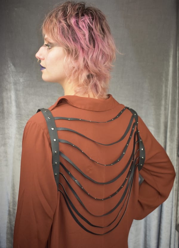 Recyled Jewellery: A woman with pink hair, viewed from behind, wearing a brown shirt with a unique back design featuring horizontal straps and eco-friendly jewelry studs, standing against a gray backdrop of Celina Harness. @ Reblack Shop