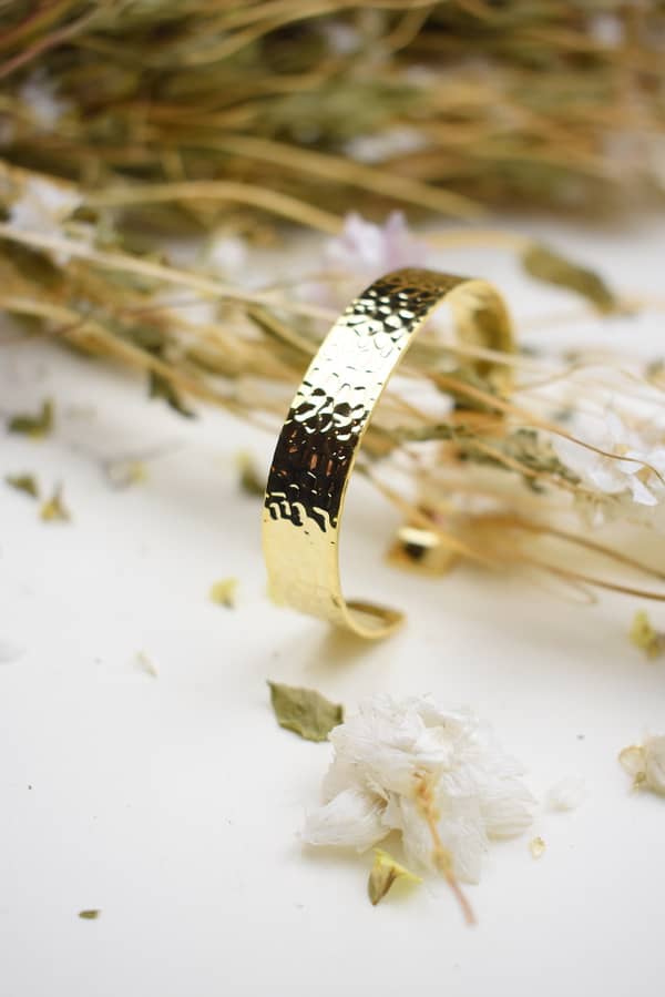 Recyled Jewellery: A golden bracelet crafted from recycled jewellery with a hammered texture, surrounded by dried flowers and petals on a white surface. @ Reblack Shop