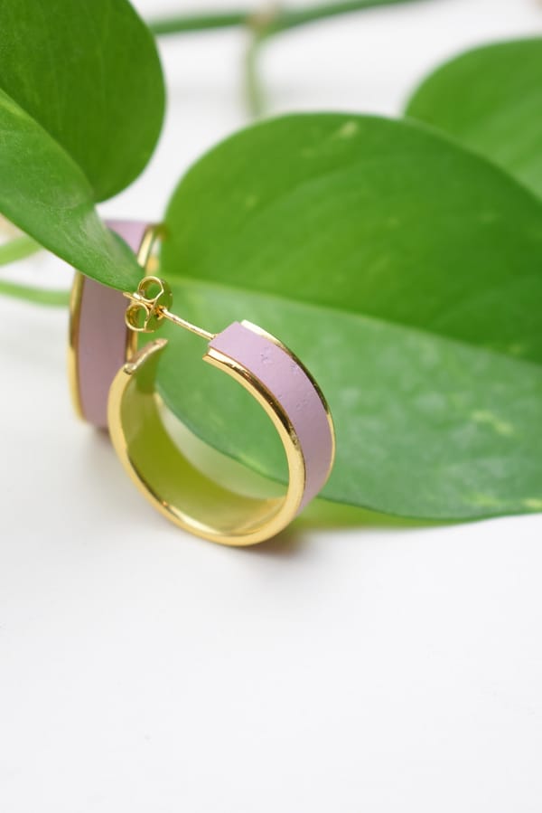 Recyled Jewellery: A close-up of a pair of recycled gold and lavender hoop earrings, artistically placed among green, oval-shaped leaves on a white background. @ Reblack Shop