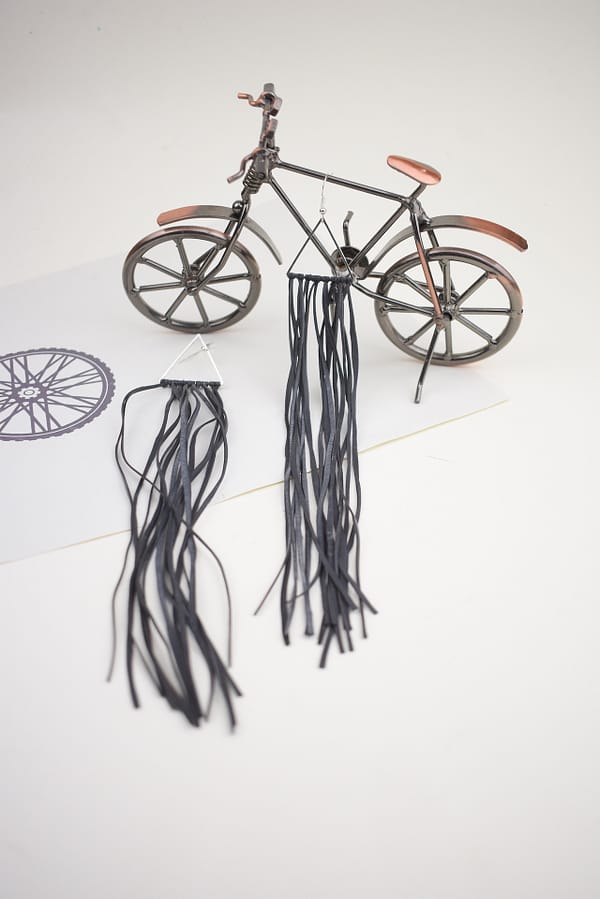 Recyled Jewellery: A miniature bicycle model crafted from recycled metal wire and leather, featuring detailed elements like handlebars and pedals, with long, black shoelace-like strands extending from the rear axle. @ Reblack Shop