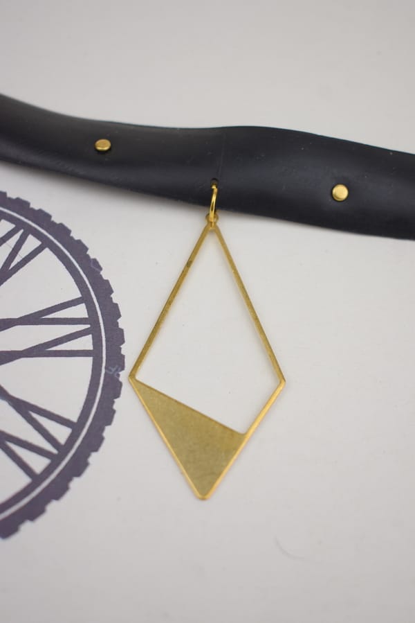 Recyled Jewellery: A close-up of a geometric gold earring made from recycled jewellery, in the shape of an inverted triangle with a solid bottom, hanging against a white background with a partial view of a bicycle wheel graphic @ Reblack Shop