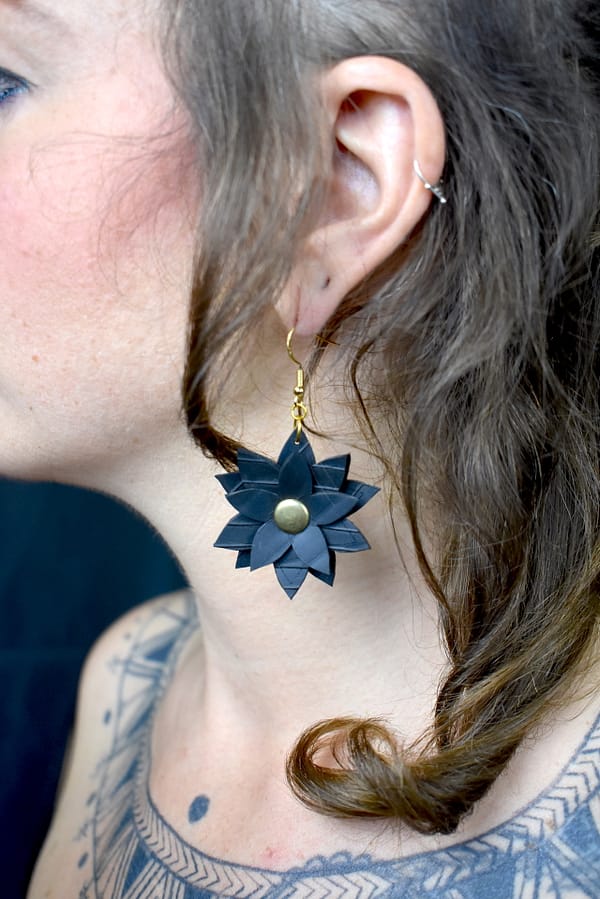 Recyled Jewellery: Close-up of a woman’s ear wearing an eco-friendly black floral earring with a gold center, set against her curly brown hair and part of a tattoo visible on her neck. @ Reblack Shop