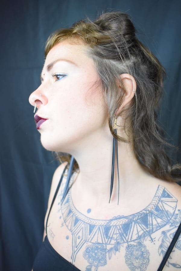 Recyled Jewellery: Side profile of a woman with a detailed chest tattoo, wearing eco-friendly jewelry earrings, against a black background. She has styled hair and dark lipstick. @ Reblack Shop