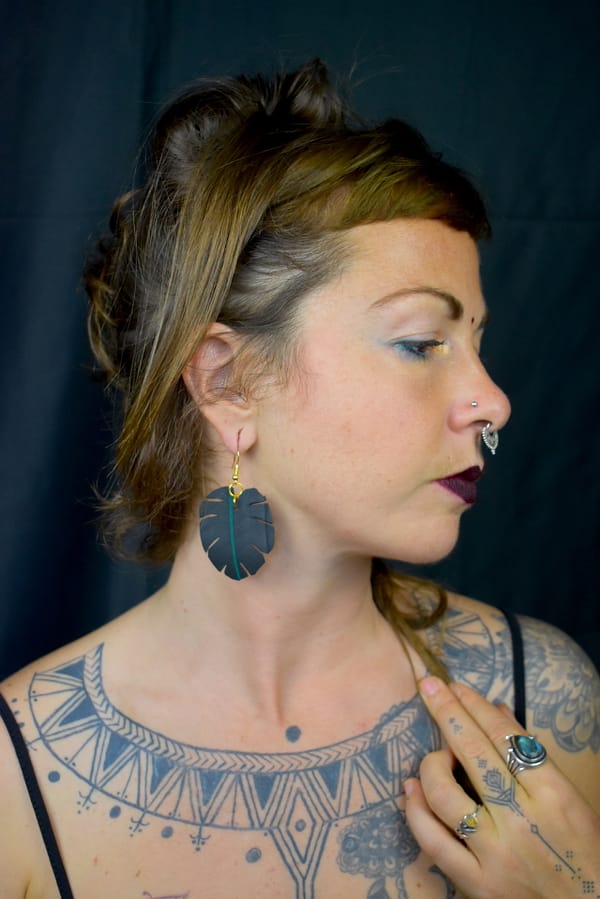 Recyled Jewellery: A woman with unique tattoos on her chest and arms, wearing a large leaf-shaped eco-friendly earring, and displaying a contemplative expression, set against a dark background. @ Reblack Shop