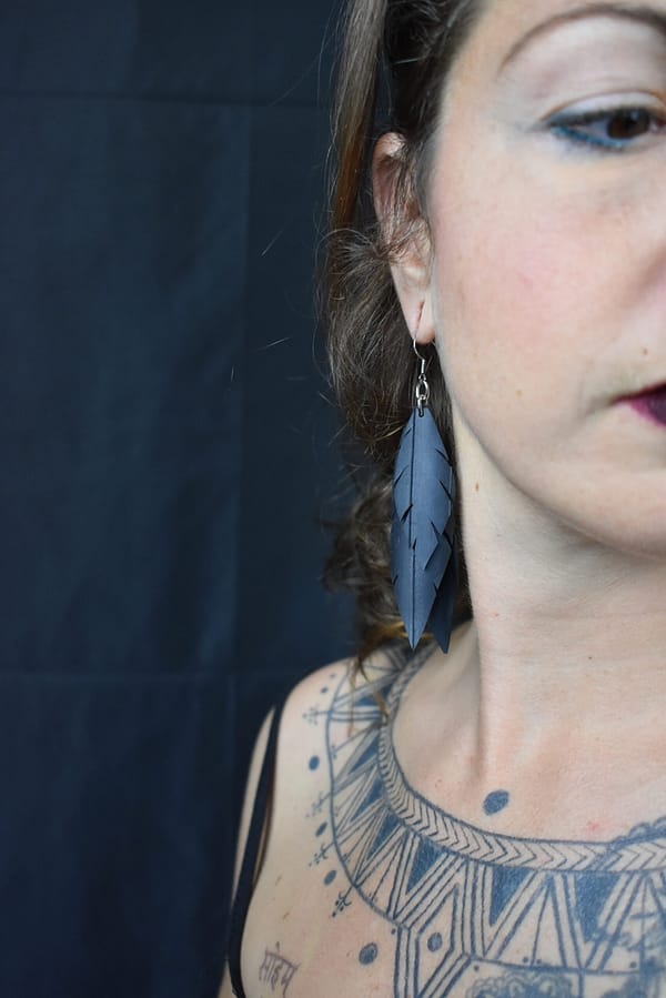 Recyled Jewellery: Close-up of a woman adorned with an eco-friendly jewelry feather earring and visible detailed tattoo art on her shoulder and neck against a dark background. @ Reblack Shop