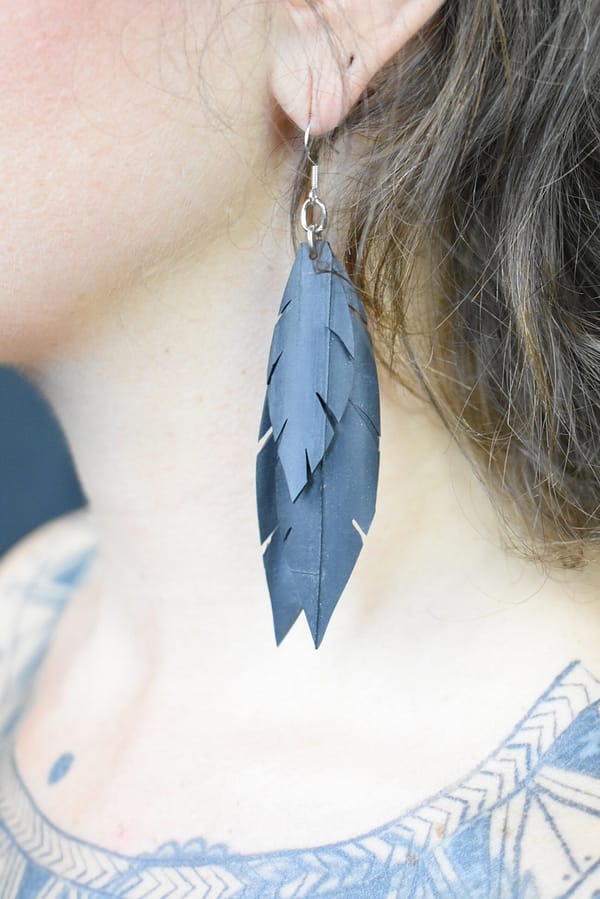 Recyled Jewellery: A close-up side view of a woman's ear wearing a long, feather-shaped earring made from repurposing jewelry, with a section of her tattooed shoulder visible. @ Reblack Shop