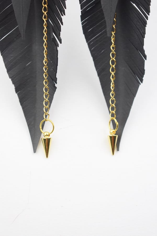 Recyled Jewellery: A pair of eco-friendly Feather Cut Golden Arrow earrings featuring gold chains and cone-shaped pendants, hanging from black feather shapes against a white background. @ Reblack Shop