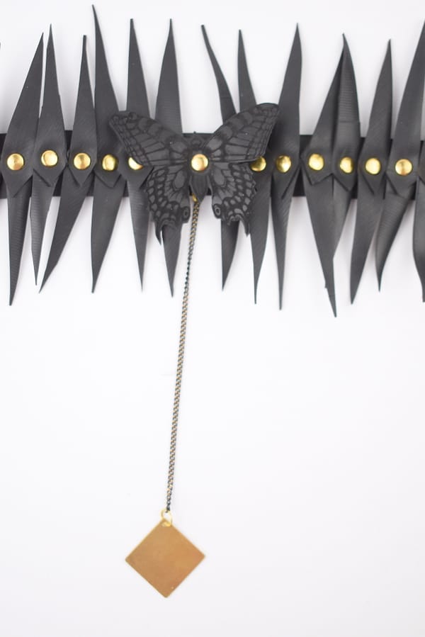 Recyled Jewellery: An artistic wall decoration featuring a series of vertical black paper leaves with gold buttons, centered by a stylized BerlinWinter Butterfly Choker with gold eyes and an eco-friendly jewelry pendant hanging below. @ Reblack Shop