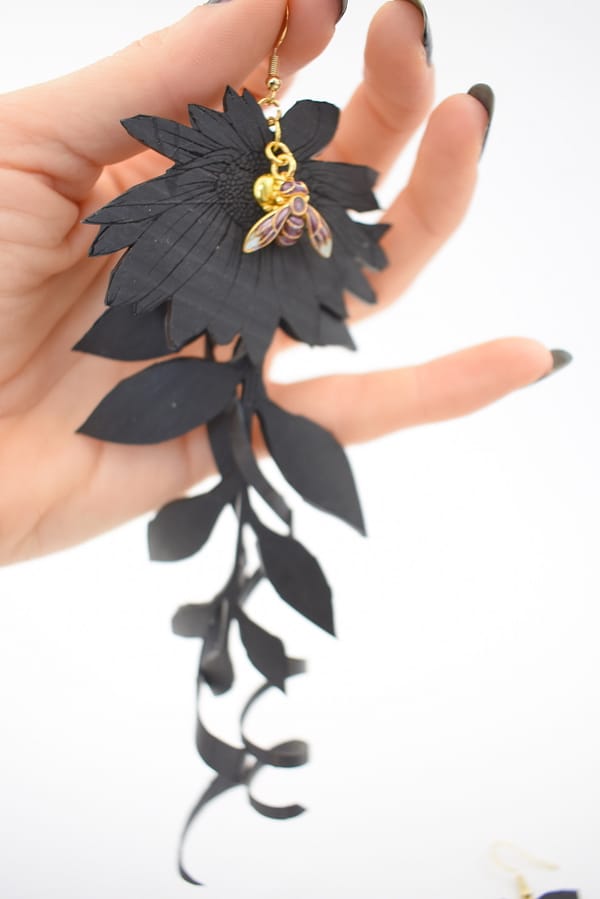 Recyled Jewellery: A hand holding the Pollinating Flowers earring with a gold and colorful bee accent against a white background. The eco-friendly earring features detailed petal and leaf designs. @ Reblack Shop